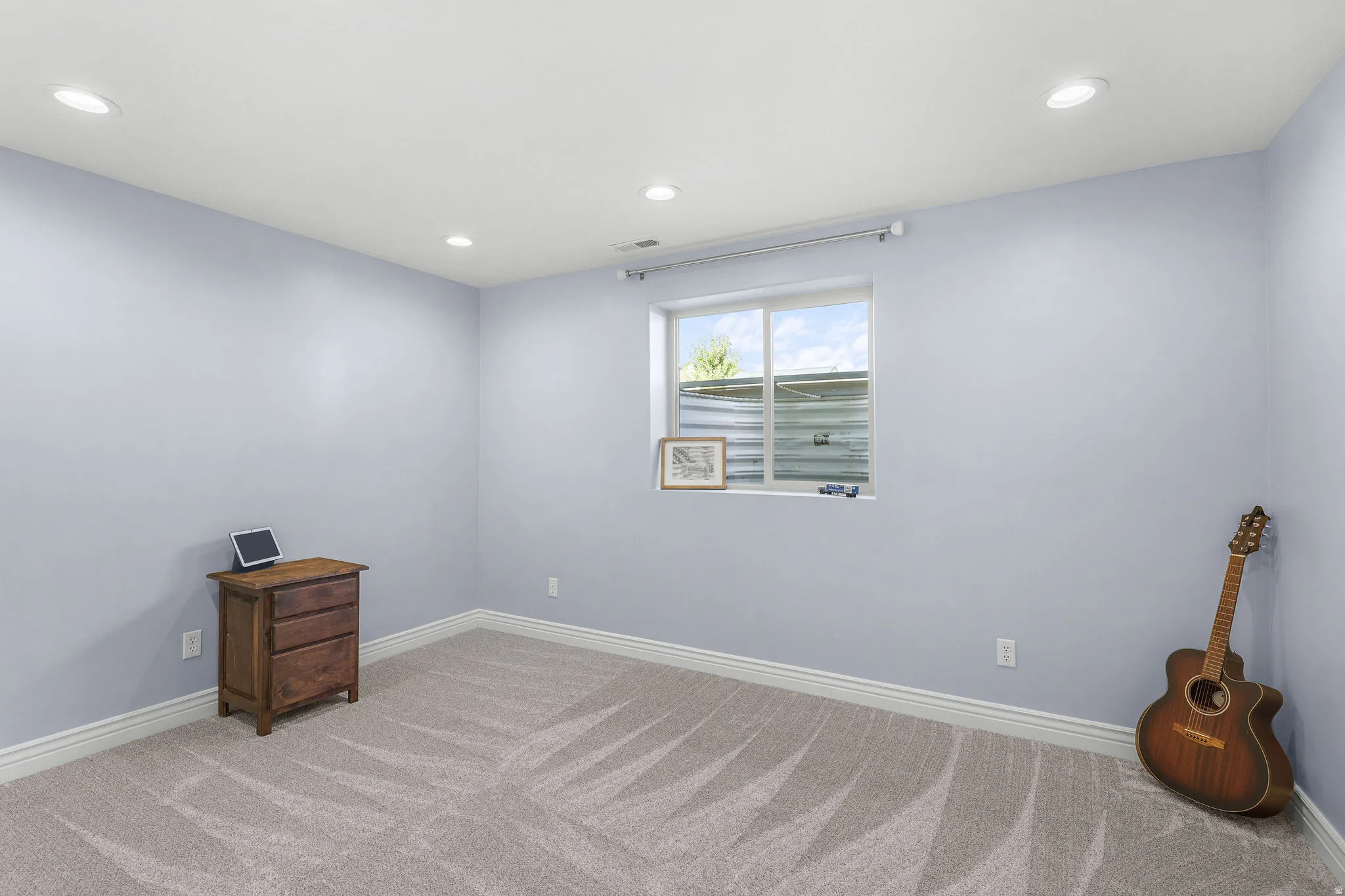 Recessed ceiling lighting, light blue wall paint, window with white trim, light gray patterned carpet, and white baseboards