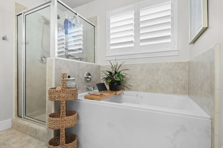 Built-in soaking tub with surrounding tile, separate glass-enclosed shower, and window with plantation shutters