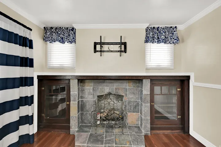 Stone-tiled fireplace with wrought iron details, flanked by two built-in wood-finish cabinets with glass doors, and two windows featuring horizontal blinds