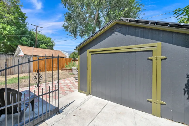 Detached storage shed with a panel door, concrete patio, paver pathway, and wrought iron gate
