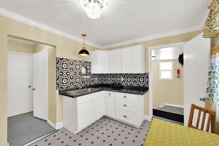 White cabinetry, dark granite-style countertops, mosaic tile backsplash, stainless steel sink, and decorative pendant lighting