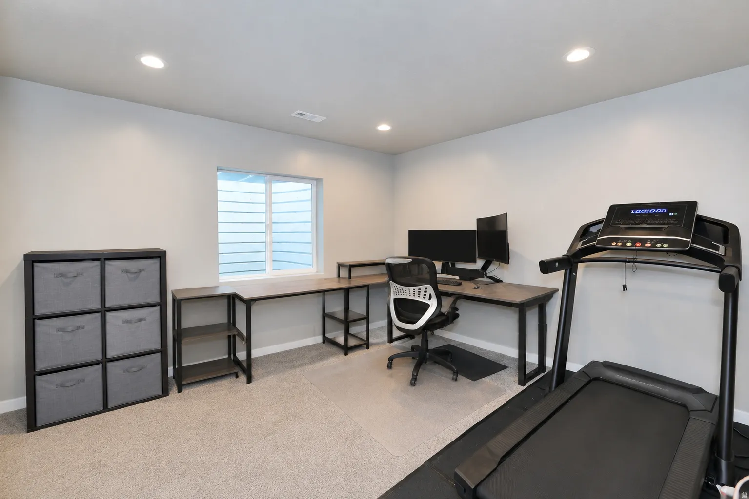 Basement bedroom currently being used as an office. Recessed lighting illuminates the space, featuring light gray walls and carpet flooring