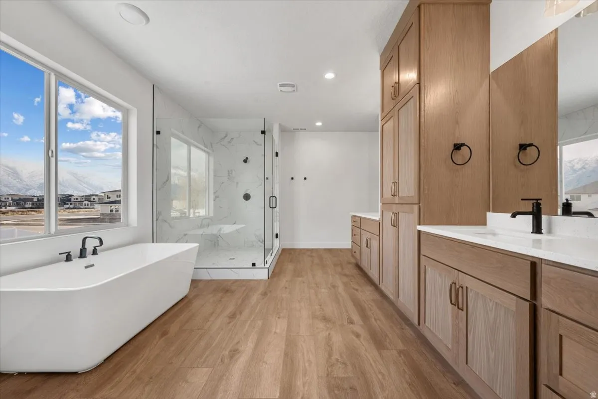 Freestanding soaking tub with matte black fixtures, glass-enclosed shower with tiled walls, wood-finish flooring, and dual vanity with light wood cabinetry and white countertops