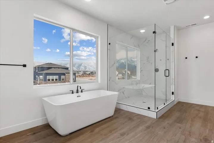 Freestanding bathtub with matte black fixtures, large window, glass-enclosed shower, white marble-style shower surround, and wood-finish flooring
