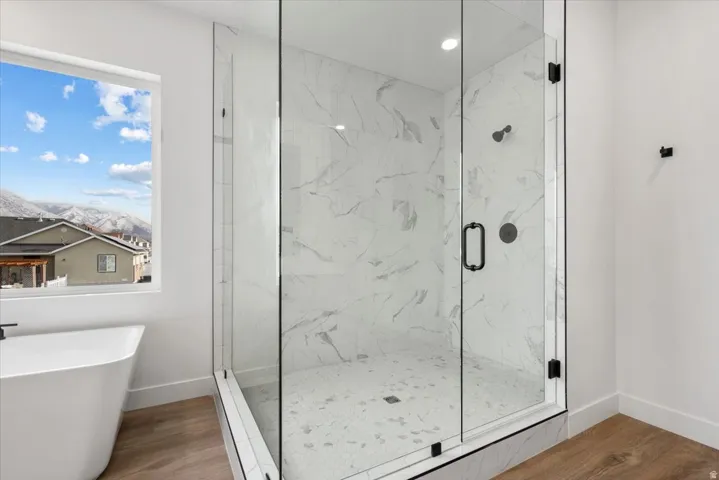 Freestanding soaking tub, glass-enclosed shower with tiled walls and floor, and wood-finish flooring