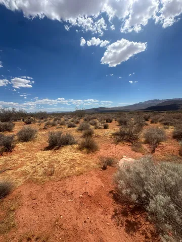 Expansive desert landscape featuring red soil, sparse brush, and distant mountains under a blue sky with clouds