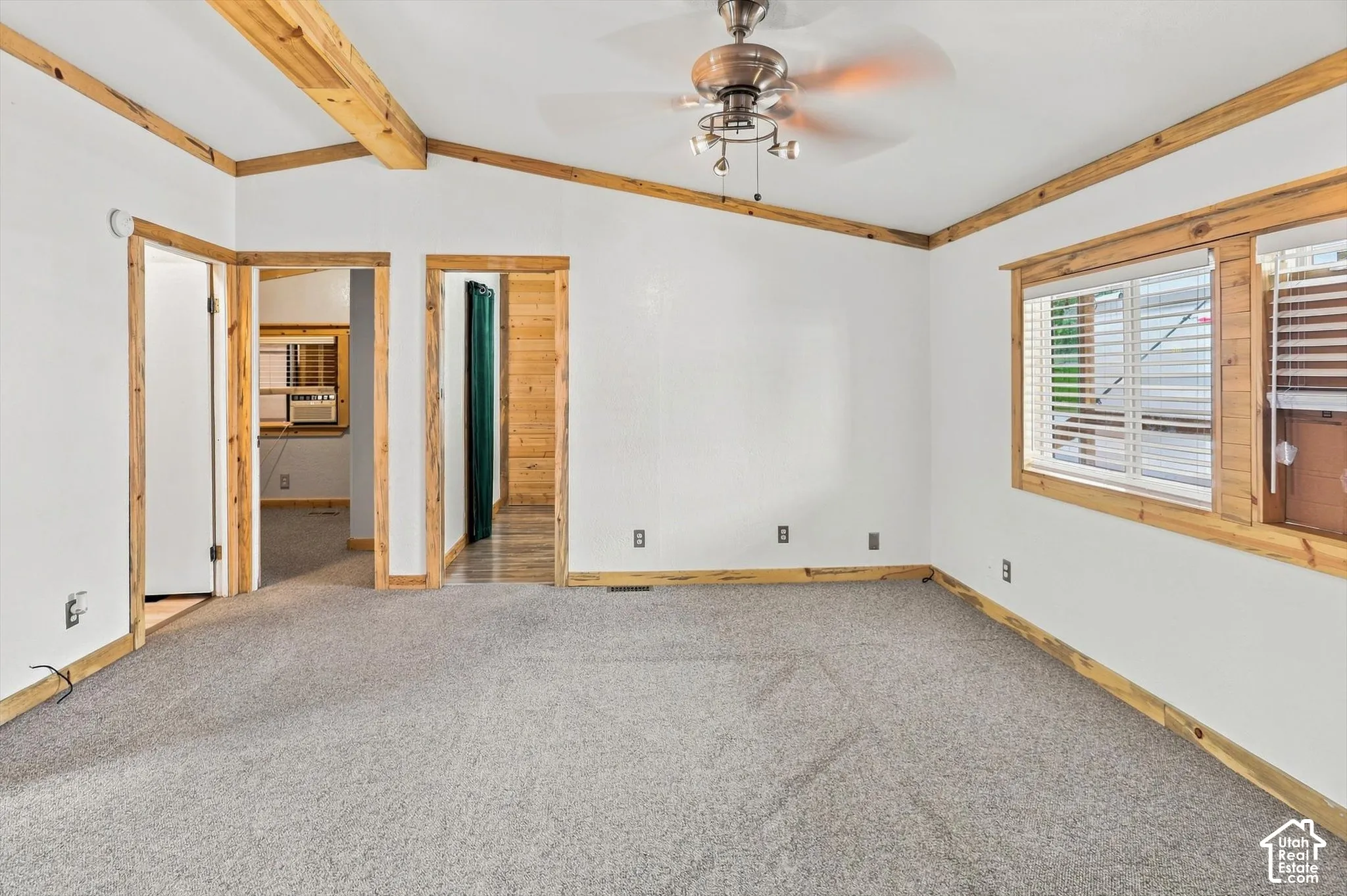 Vaulted ceiling with exposed wood beams, ceiling fan with integrated lighting, wood-trimmed windows with horizontal blinds, and wall-to-wall carpeting
