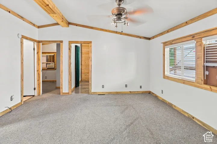 Vaulted ceiling with exposed wood beams, ceiling fan with integrated lighting, wood-trimmed windows with horizontal blinds, and wall-to-wall carpeting