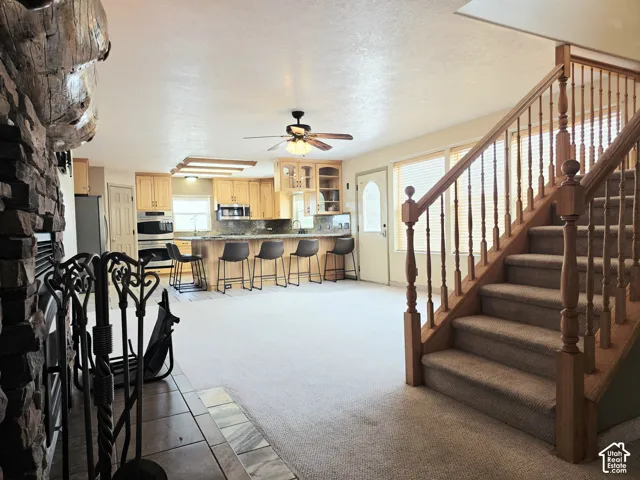 Spacious open-concept living area featuring a stone fireplace, wood staircase, wood-finish cabinetry, stainless steel appliances, and a ceiling fan