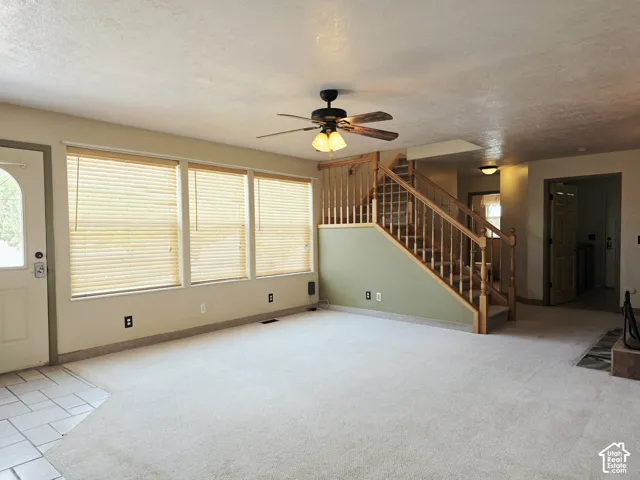 Carpeted living area with a wood staircase, three windows with blinds, a ceiling fan, and tile flooring at the entry