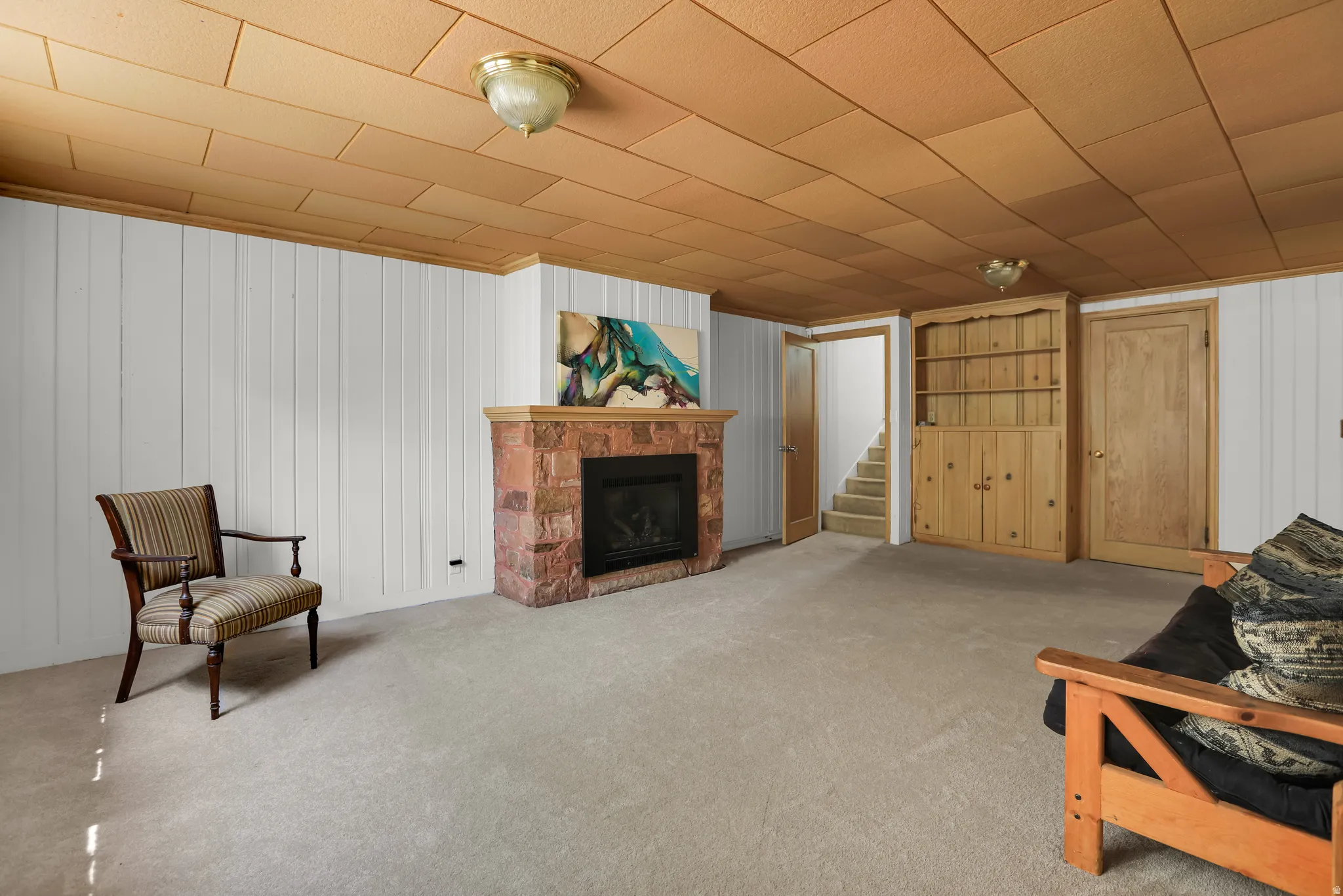 Spacious basement recreation room featuring a brick fireplace with hearth, wood-finish ceiling with recessed lighting, neutral carpet flooring, and multiple wooden storage closets