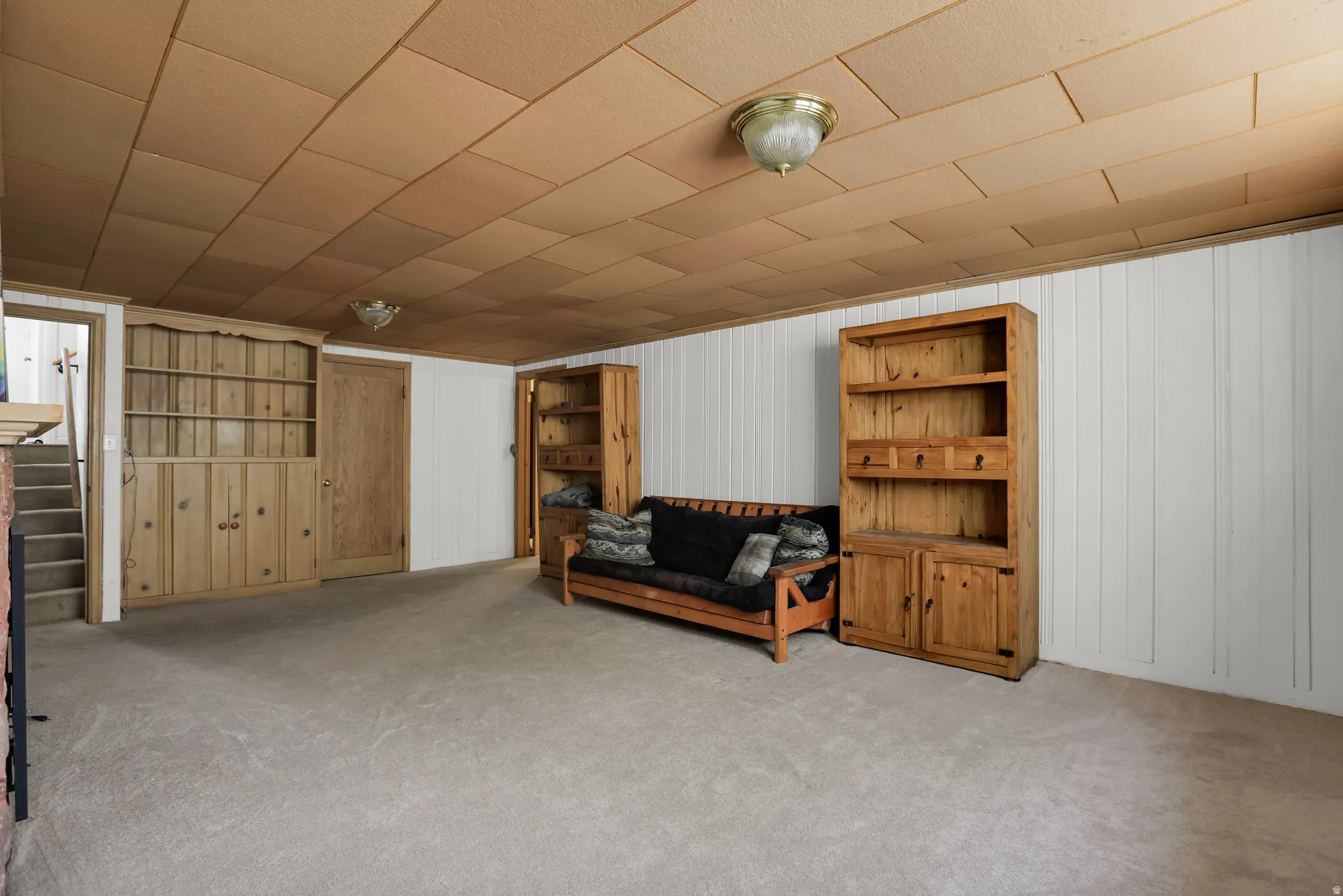 Carpeted room featuring a built-in wood-finish cabinet with shelving, additional wood-finish shelving unit, white paneling, and a panel-style ceiling