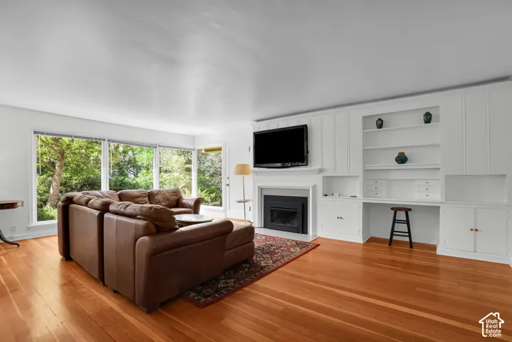 Hardwood flooring throughout a living space featuring a fireplace with a white mantel, an integrated media center, built-in shelving, and a desk area