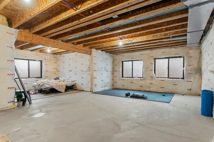 Unfinished basement space with exposed wooden beams, concrete flooring, and multiple windows