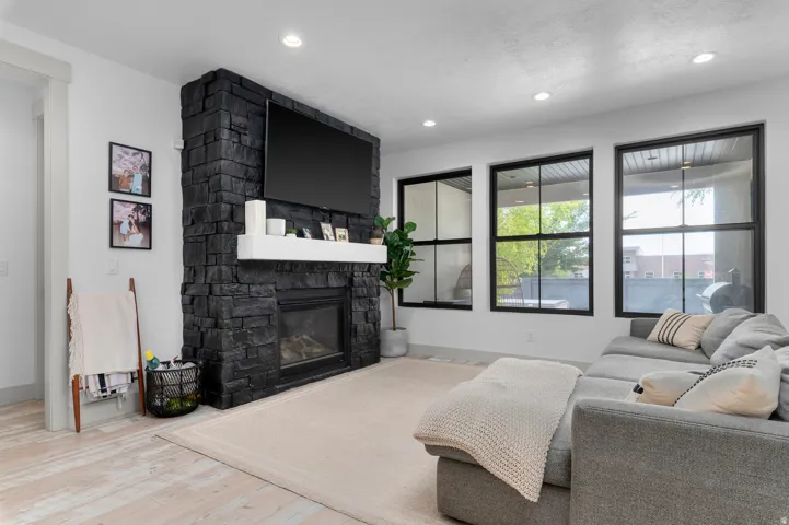 Floor-to-ceiling black stone fireplace with a white mantel, wide plank wood-finish flooring, recessed lighting, and large black-framed windows