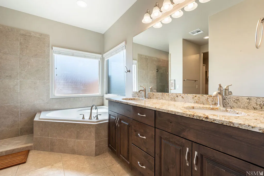 Dual sink vanity with granite countertop and dark wood-finish cabinetry, complemented by a built-in corner soaking tub and tiled walls