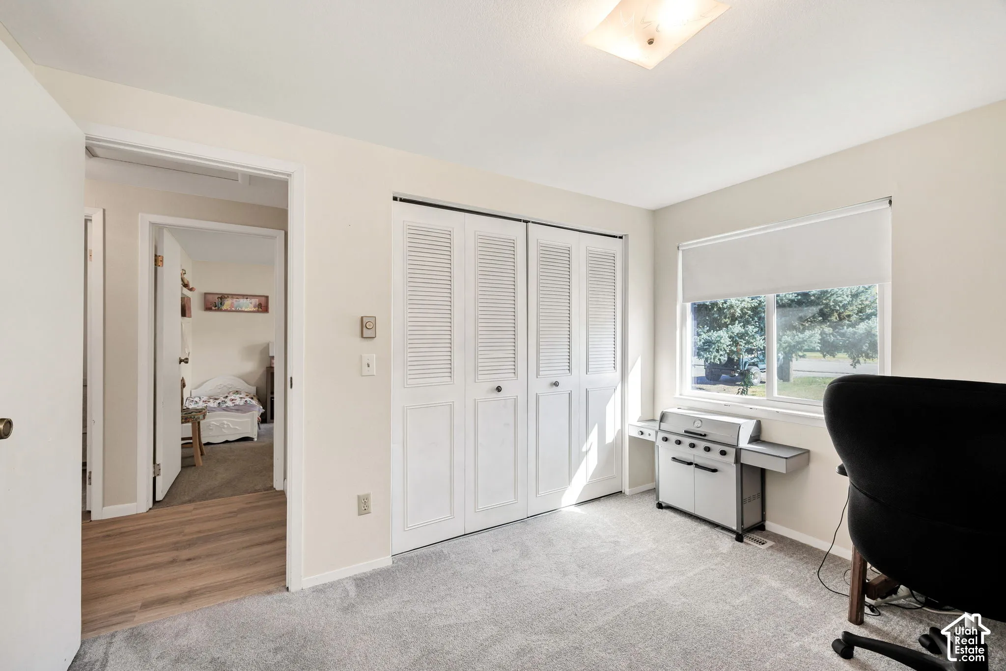 Carpeted room featuring a window with roller shade, louvered bi-fold closet doors, and overhead lighting