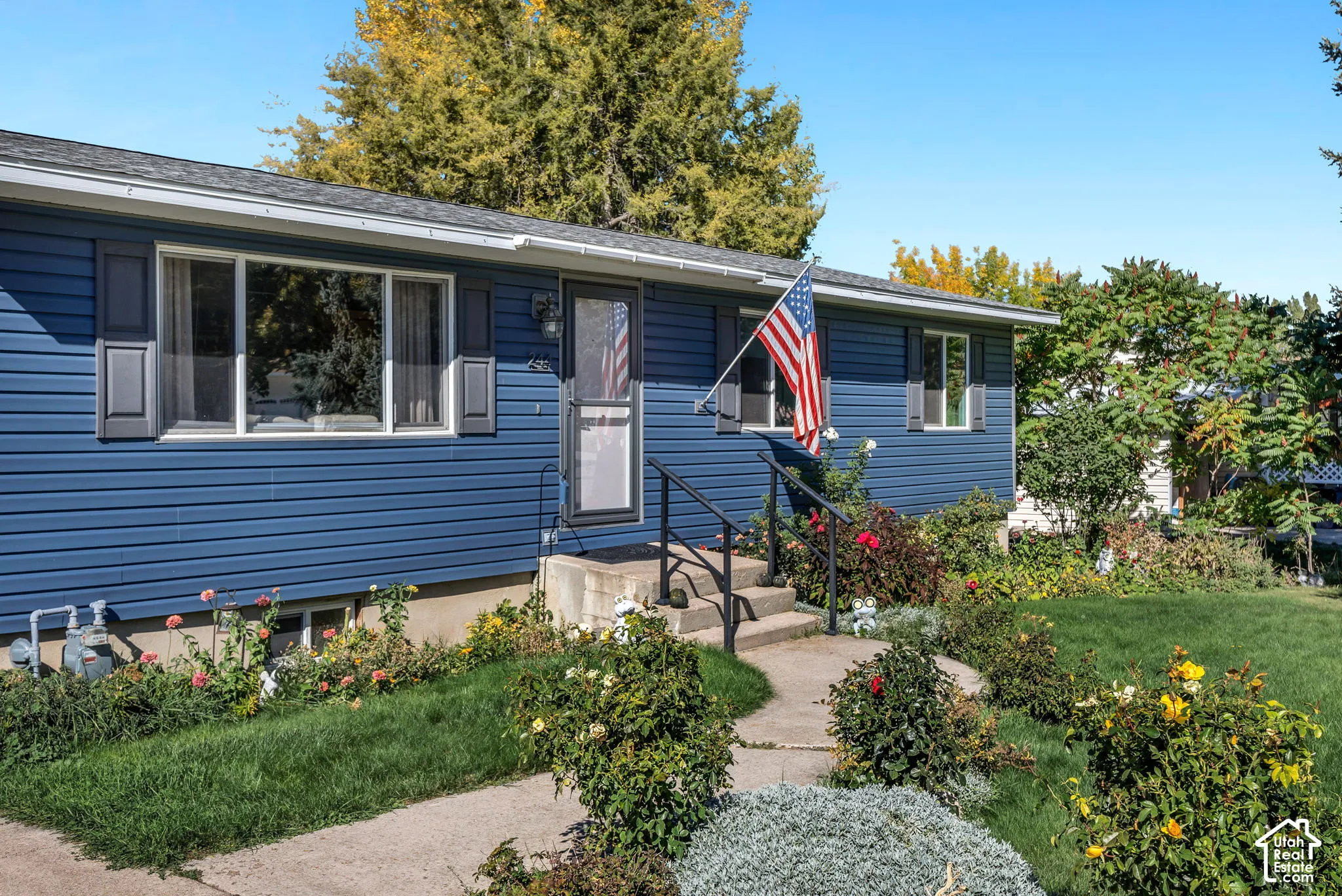 Blue vinyl siding exterior, concrete steps with metal railing, front entry storm door, large picture window with dark shutters, and established landscaping
