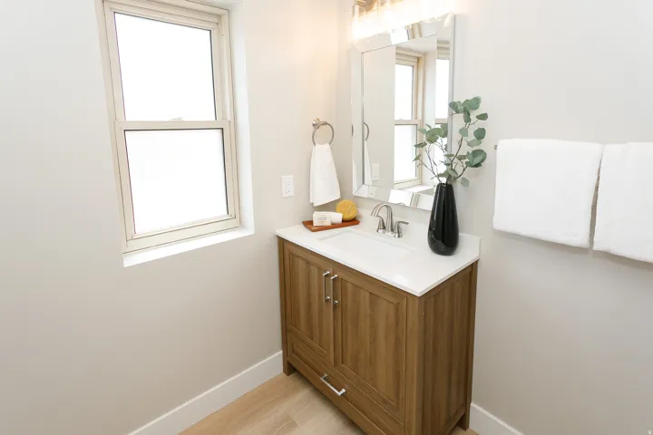 Wood-finish vanity with an integrated sink and brushed nickel faucet, framed mirror with overhead lighting, and a single window