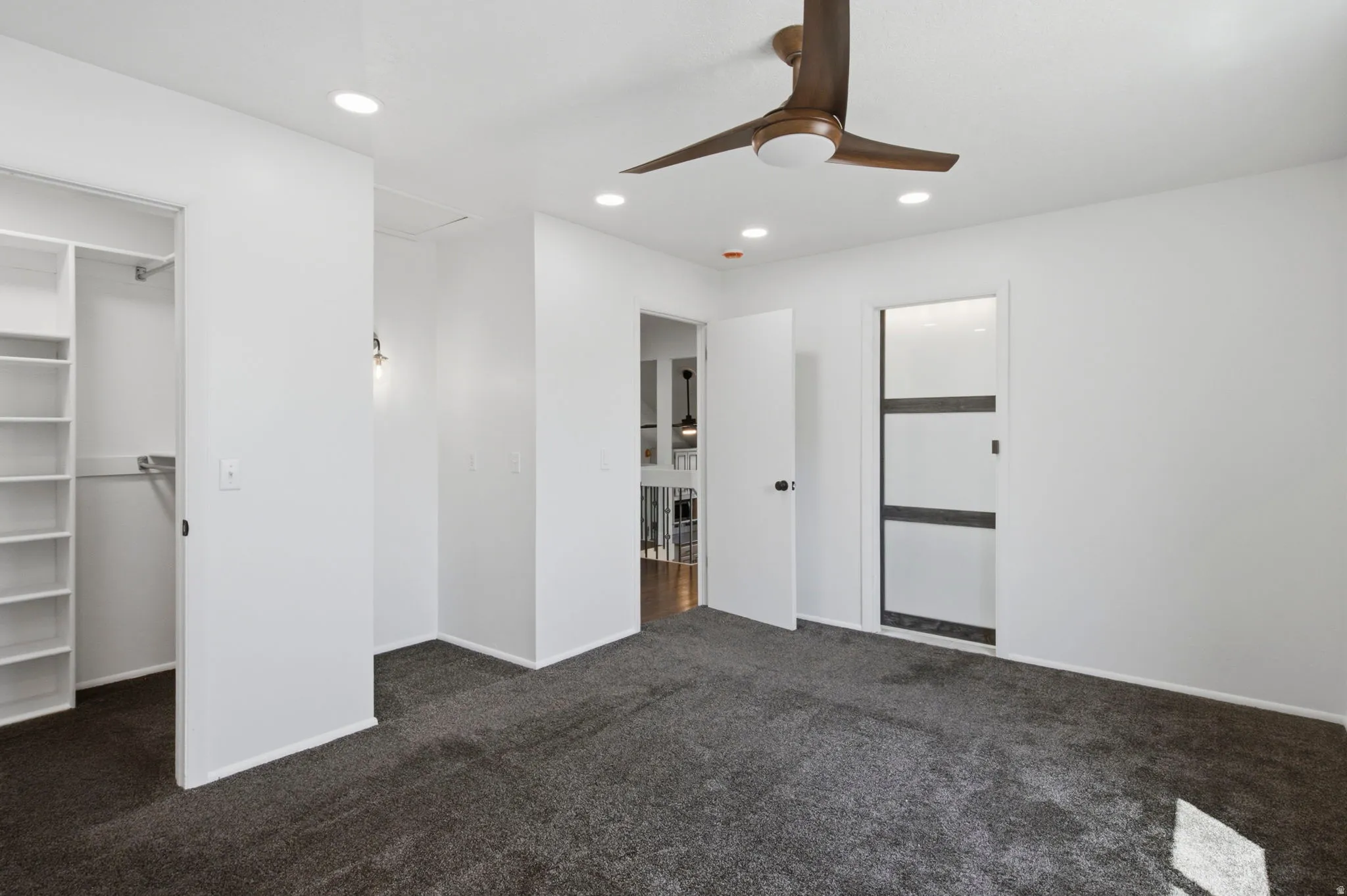 Recessed lighting, ceiling fan, built-in shelving, gray carpeting, and a modern interior door with frosted panels