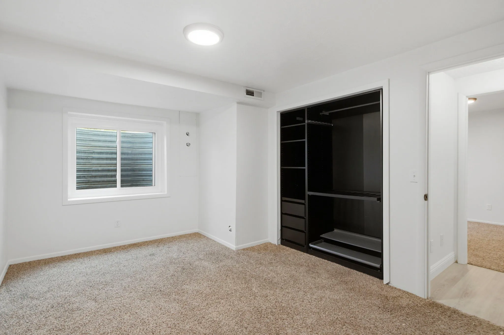 Spacious bedroom with neutral carpet flooring, large walk-in closet featuring black framed glass sliding doors and built-in shelving, recessed ceiling lighting, and a window with plantation shutters providing natural light