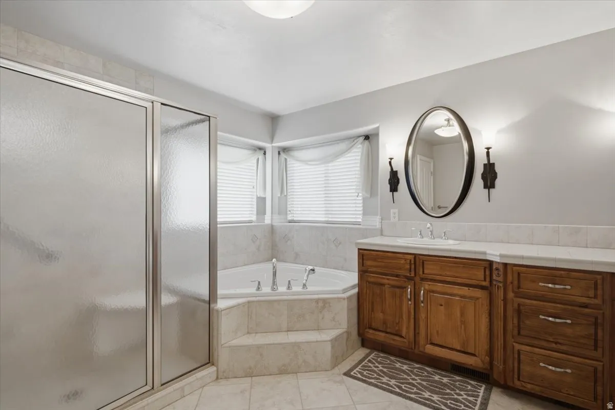 Tiled walk-in shower with frosted glass enclosure, corner soaking tub with tiled surround, dual-sink vanity with wood-finish cabinetry, light-toned countertop, and an oval mirror with sconce lighting