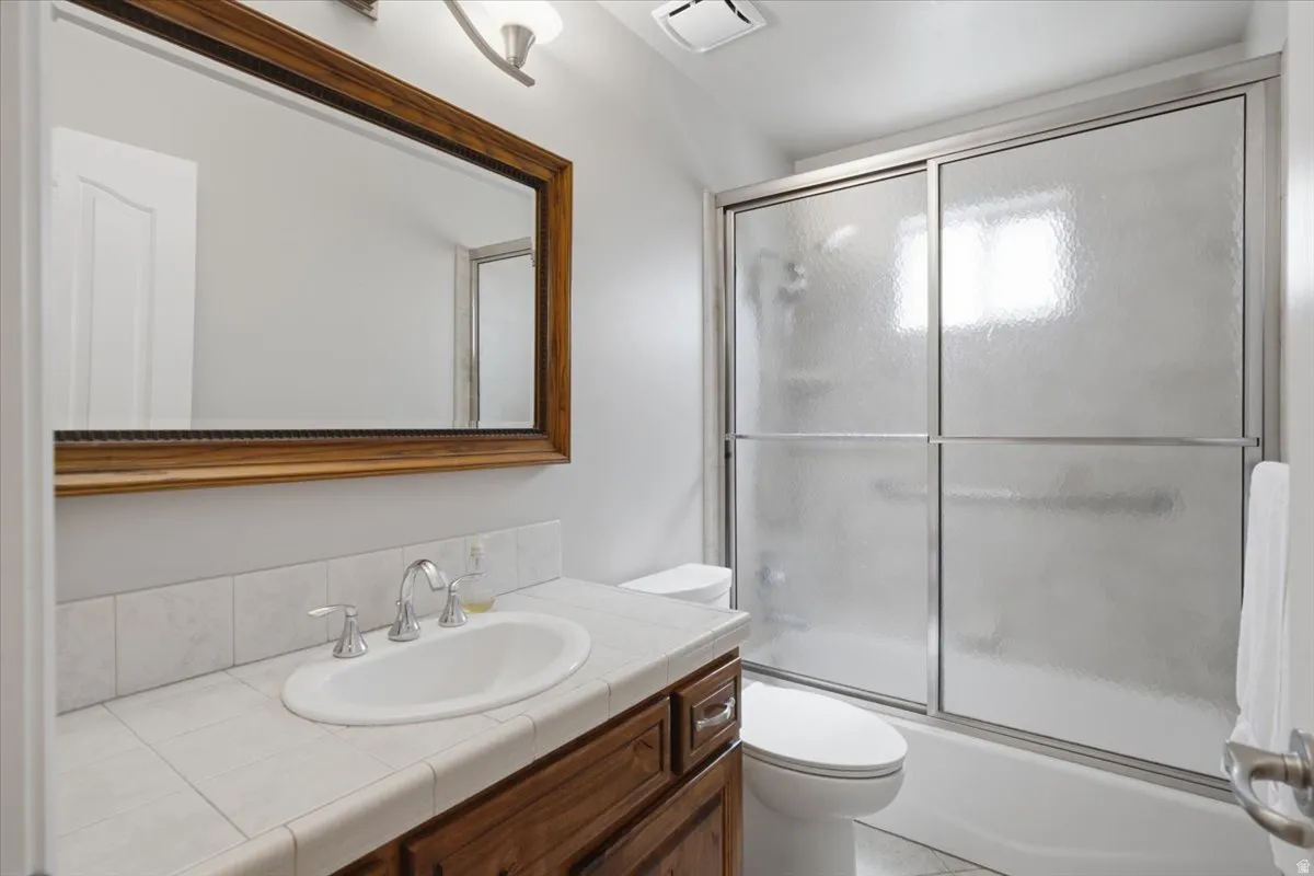 Wood-framed vanity mirror, single basin sink with chrome fixtures, tiled countertop, dark wood-finish cabinetry, and a shower-tub combination with sliding obscured glass doors