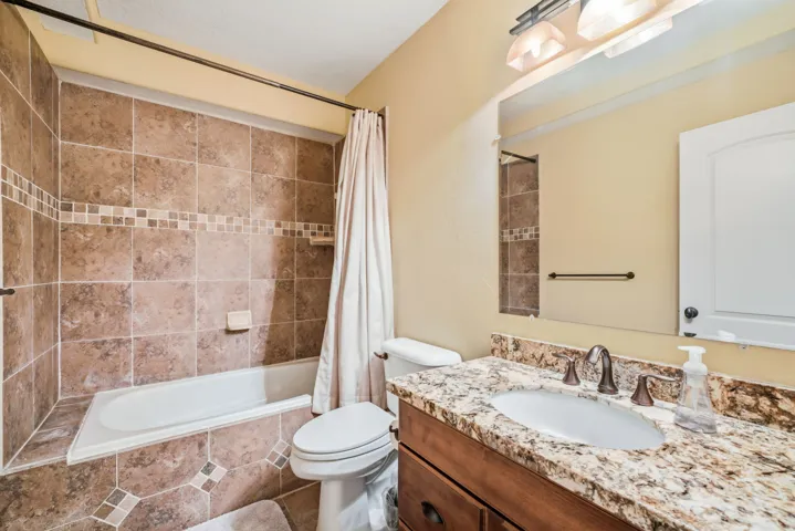 Tile-surround bathtub, tiled flooring, vanity with a rectangular sink, stone-finish countertop, and wall-mounted light fixture