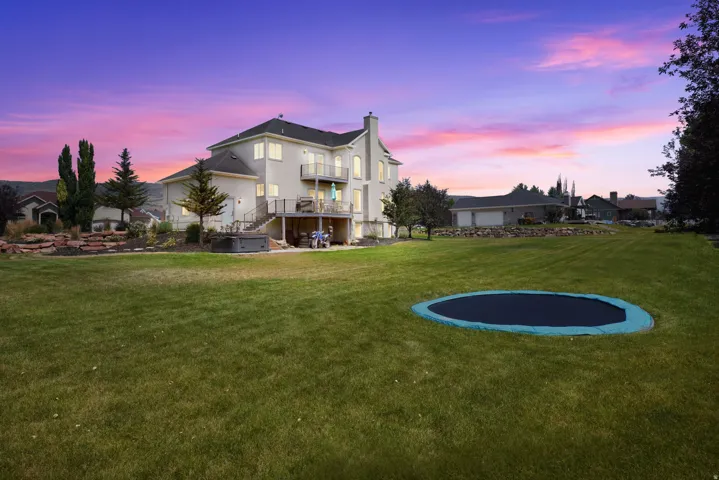 Expansive manicured lawn with mature landscaping, two-story Colonial-style residence featuring a light-colored exterior with multi-level deck construction, and in-ground trampoline with blue safety padding set against a backdrop of evergreen trees and nei