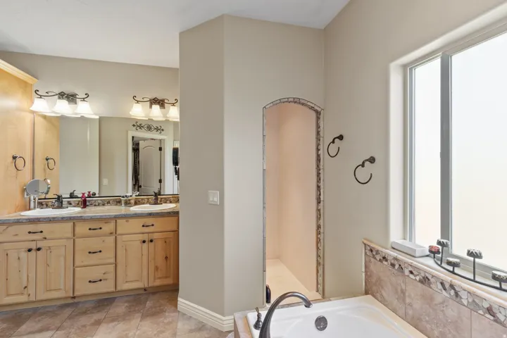 Dual sink vanity with wood-finish cabinetry, built-in soaking tub, arched alcove, and a large privacy window