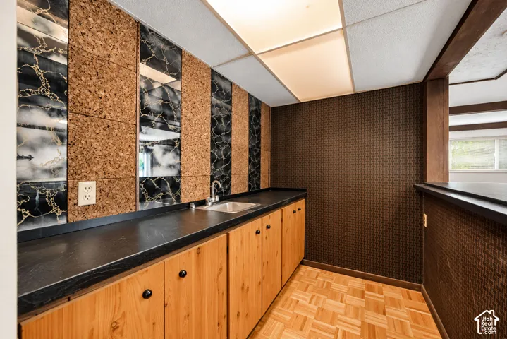 Wet bar featuring custom wood cabinetry with black countertops, cork and marble-look wall panels, recessed ceiling lighting, and warm wood-finish flooring throughout
