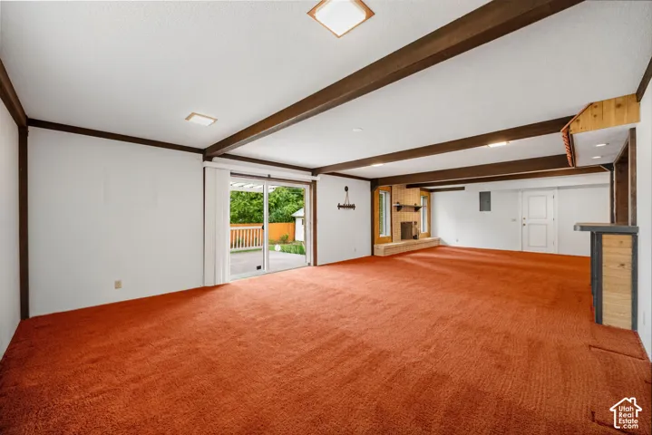 Spacious recreation room featuring exposed beam ceiling, brick fireplace with metal surround and hearth, wood-paneled accent wall with transom windows, and recessed lighting.