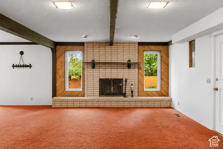 Spacious recreation room featuring exposed beam ceiling, brick fireplace with metal surround and hearth, wood-paneled accent wall with transom windows, and recessed lighting.
