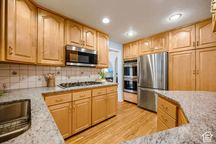 Bright galley kitchen showcasing warm wood cabinetry, stainless steel appliances including a gas cooktop and dual wall ovens, granite countertops, recessed ceiling lighting, and wood-finish flooring throughout