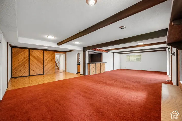 Spacious recreation room featuring exposed beam ceiling, brick fireplace with metal surround and hearth, wood-paneled accent wall with transom windows, and recessed lighting.