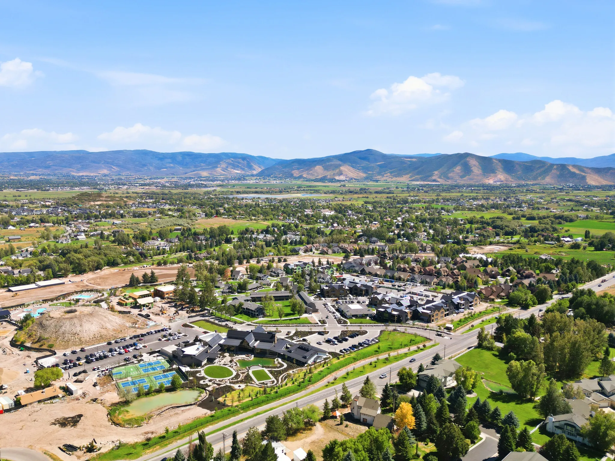 Expansive aerial perspective featuring a development with multiple structures, tennis courts, and a parking area, surrounded by lush green landscapes and distant mountains