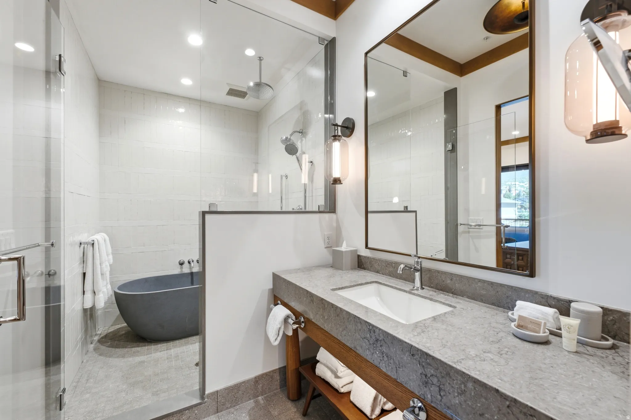 Freestanding oval bathtub, glass-enclosed shower, large rectangular vanity mirror, stone countertop with integrated sink, and exposed wood vanity shelving