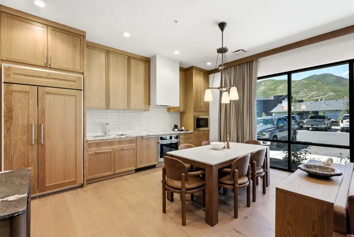 Integrated kitchen and dining area featuring wood-finish flooring, wood cabinetry, built-in panel-ready refrigerator, stainless steel appliances, and large windows