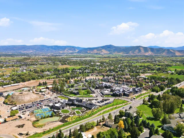 Expansive aerial perspective featuring a development with multiple structures, tennis courts, and a parking area, surrounded by lush green landscapes and distant mountains