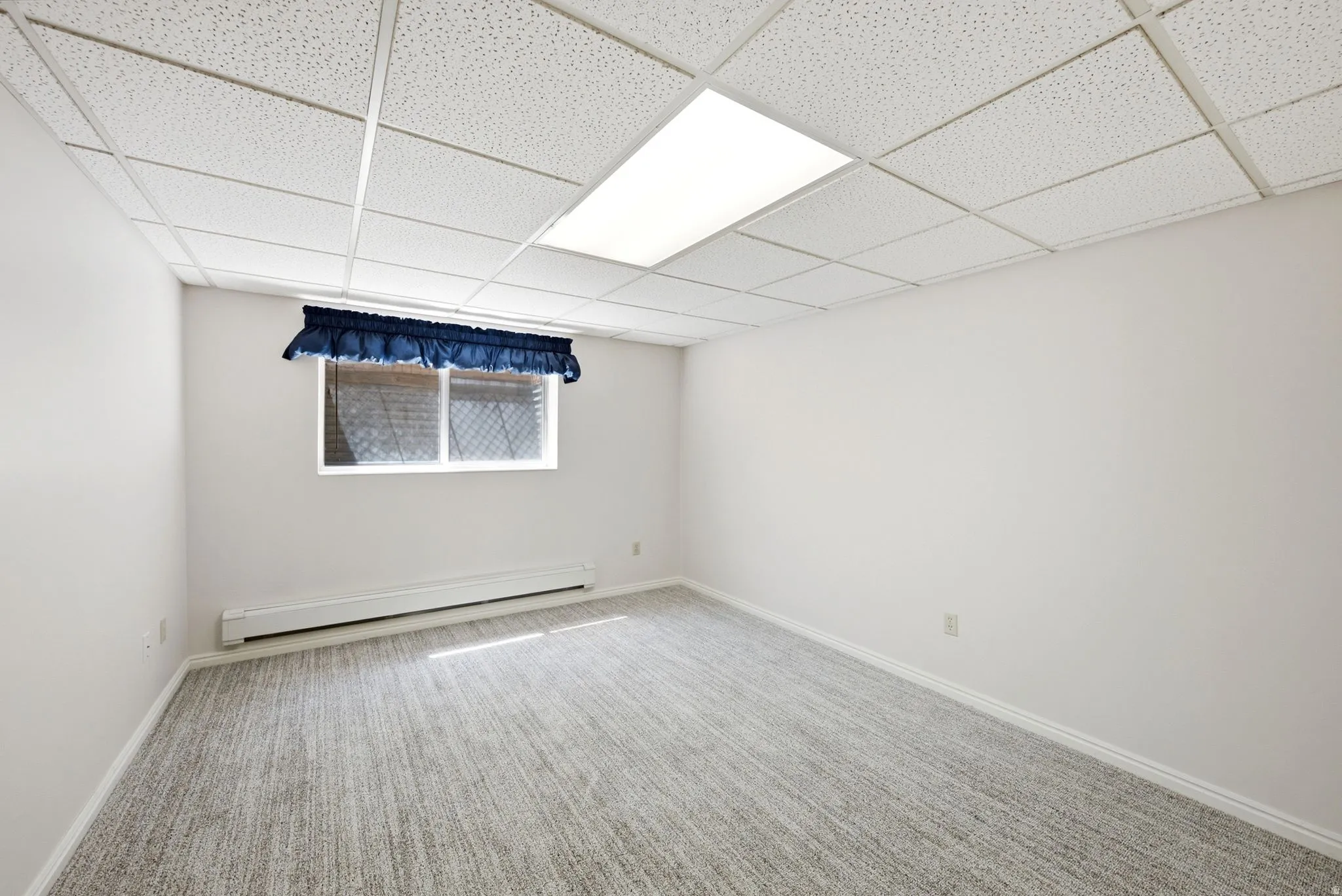 White painted walls, textured carpet flooring, window with blue valance, baseboard heating unit, and suspended ceiling with integrated lighting