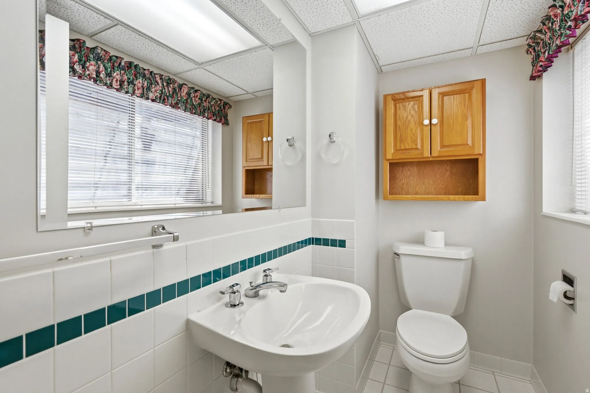 Pedestal sink, white tile flooring, built-in wooden cabinetry, window with blinds, and recessed lighting