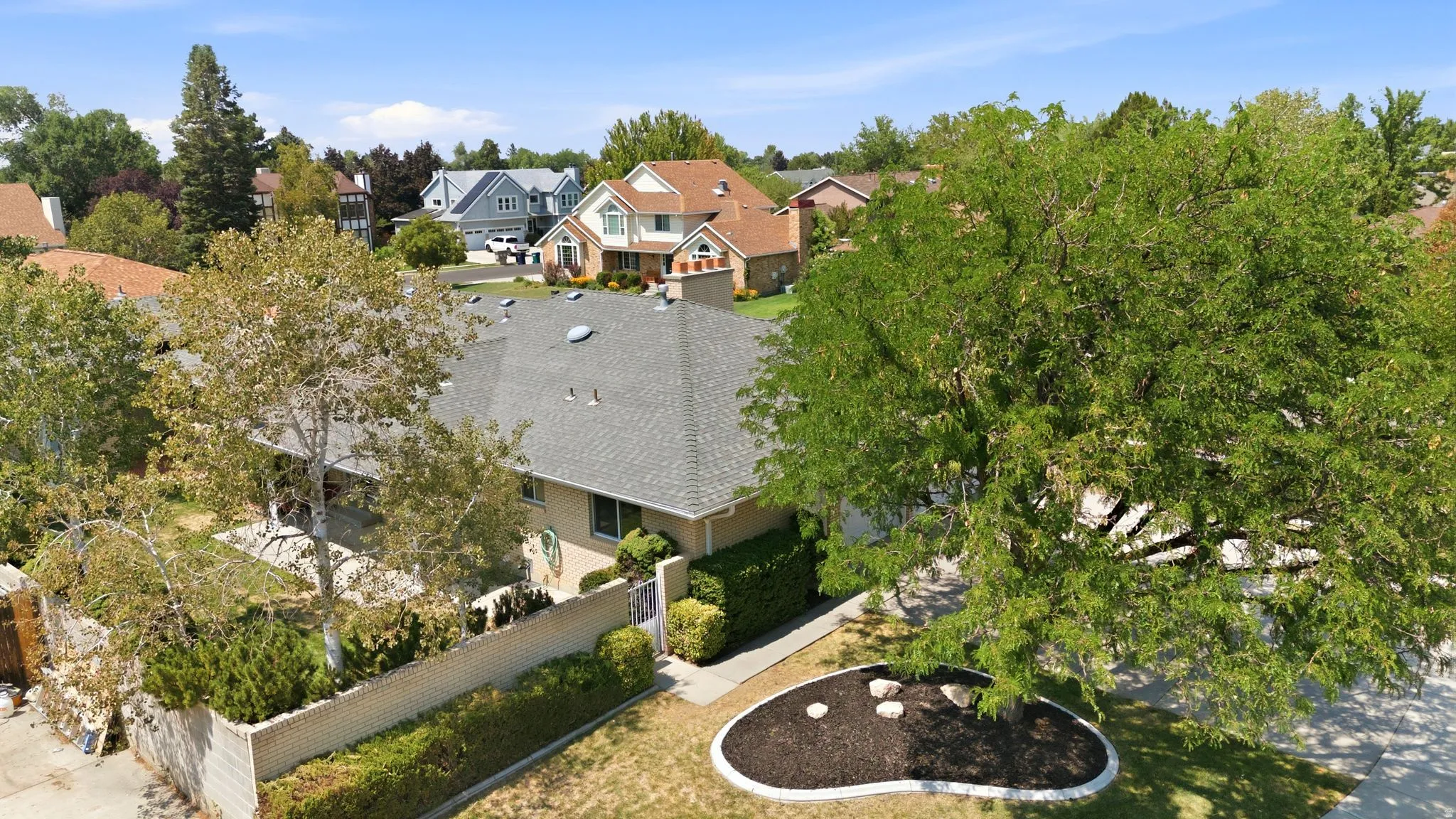 Pitched roof with gray shingles, mature perimeter trees, and a landscaped front yard with decorative rocks