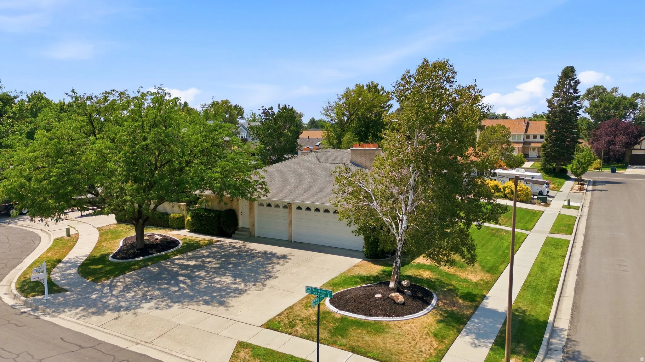 Single-story residence featuring a paved driveway, attached garage, mature front yard landscaping with shade trees, and a concrete sidewalk