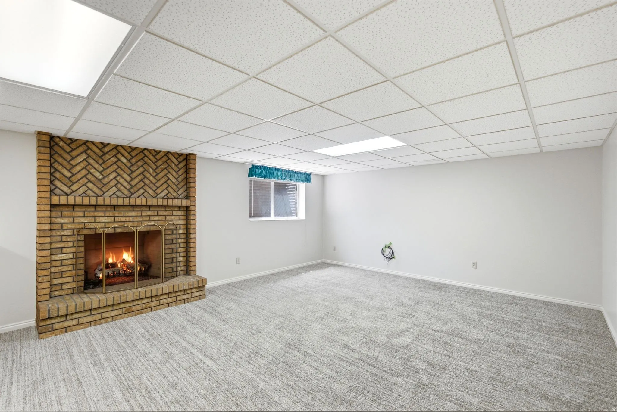 Brick fireplace with herringbone detail, drop ceiling with integrated lighting, window with blinds, and neutral carpeting
