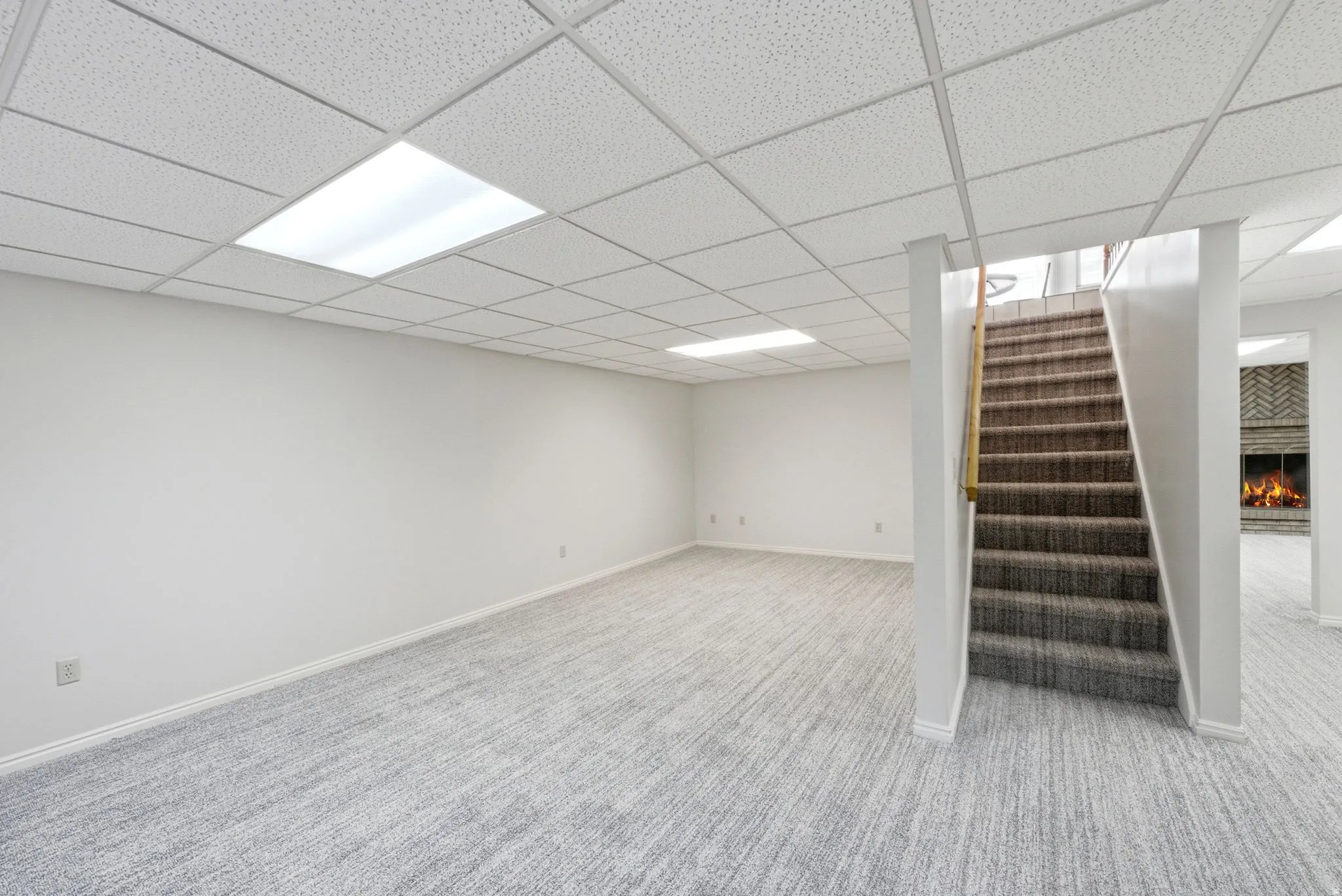 Spacious room with light-toned carpet, drop ceiling with integrated lighting, and a staircase with wood-toned handrail