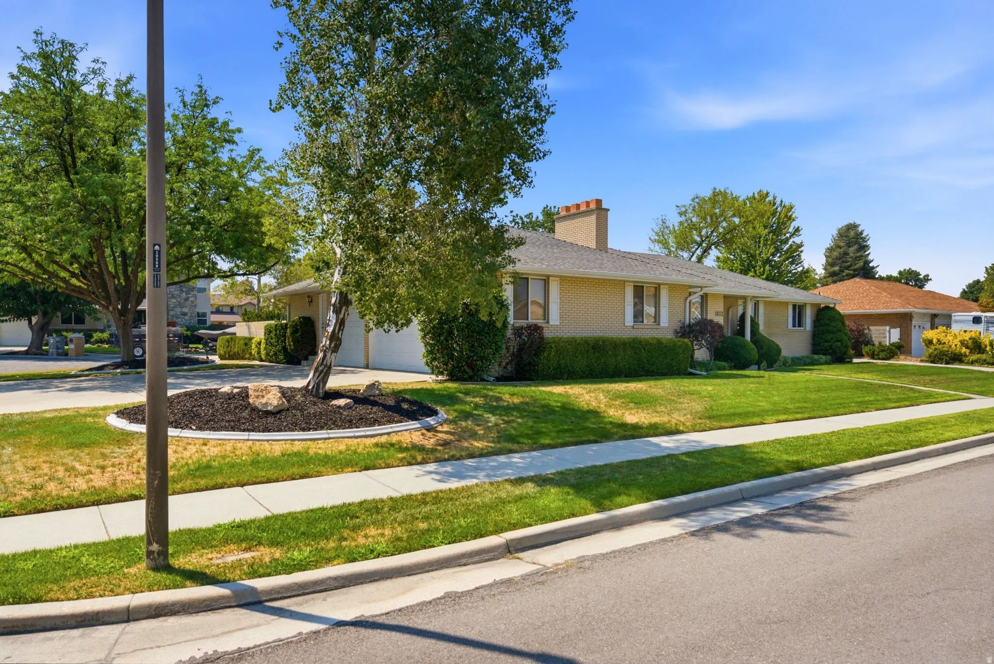 Brick exterior with a prominent chimney feature, manicured front lawn, established landscaping, and a concrete sidewalk