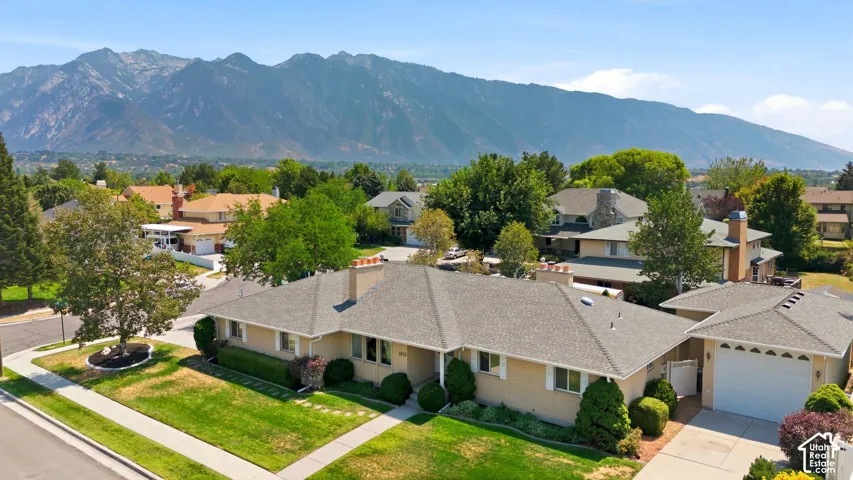 Mountain backdrop, mature landscaping, paved driveway, attached garage, and a sidewalk leading to the entrance
