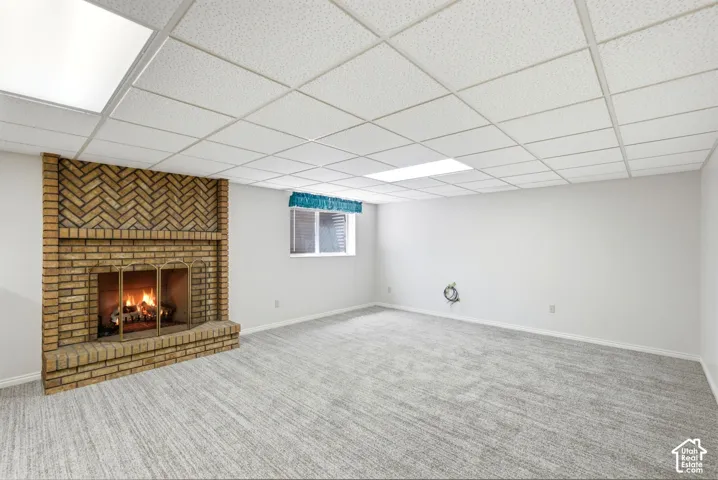 Brick fireplace with herringbone detail, drop ceiling with integrated lighting, window with blinds, and carpeting
