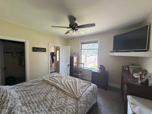 Carpeted bedroom featuring a ceiling fan with integrated lighting, a window with white trim, and a wall-mounted shelf