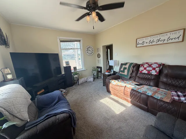 Living area featuring neutral-toned walls, light-colored carpeting, a ceiling fan with integrated lighting, and a window with architectural detailing